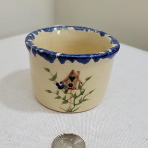 May include: A small, cream-colored ceramic bowl with a blue painted rim and a hand-painted design of a brown birdhouse on a green vine. A US quarter coin is placed in front of the bowl to provide a clear sense of scale for the object.