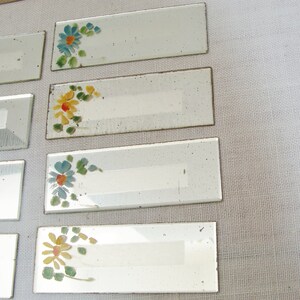 May include: Rectangular glass name tags with hand-painted floral designs in blue, yellow, and orange. The tags have a white rectangular space for writing. The tags are arranged on a light-colored surface.