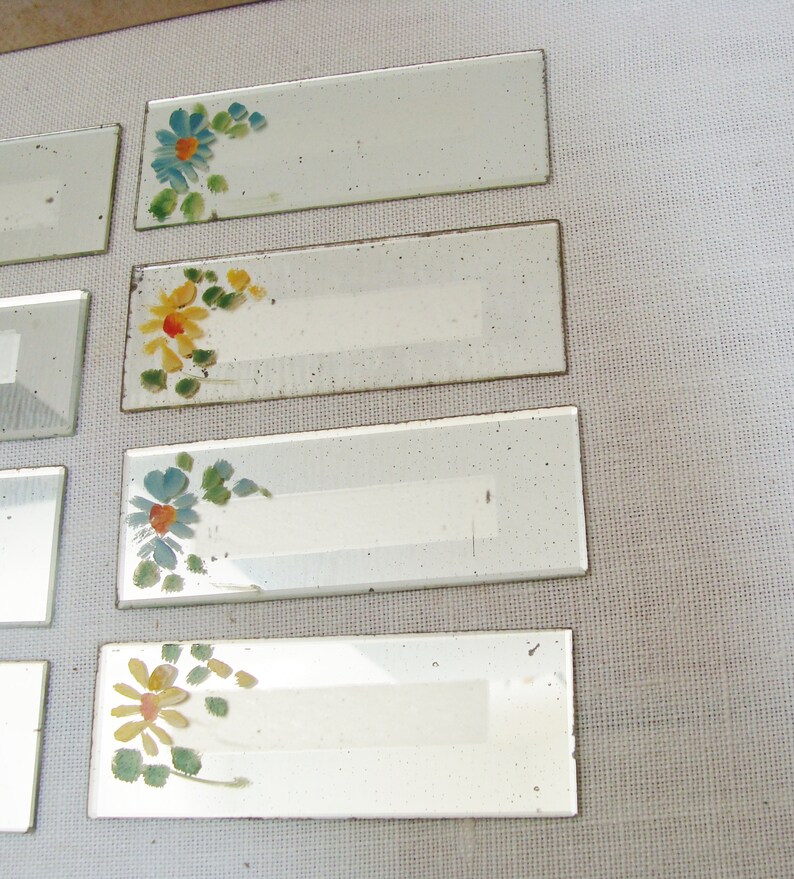 May include: Rectangular glass name tags with hand-painted floral designs in blue, yellow, and orange. The tags have a white rectangular space for writing. The tags are arranged on a light-colored surface.