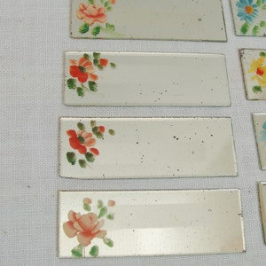 May include: Rectangular, mirrored tiles with hand-painted floral designs. The flowers are in shades of red, orange, and pink, with green leaves. The tiles are arranged on a white surface, showcasing a vintage aesthetic.