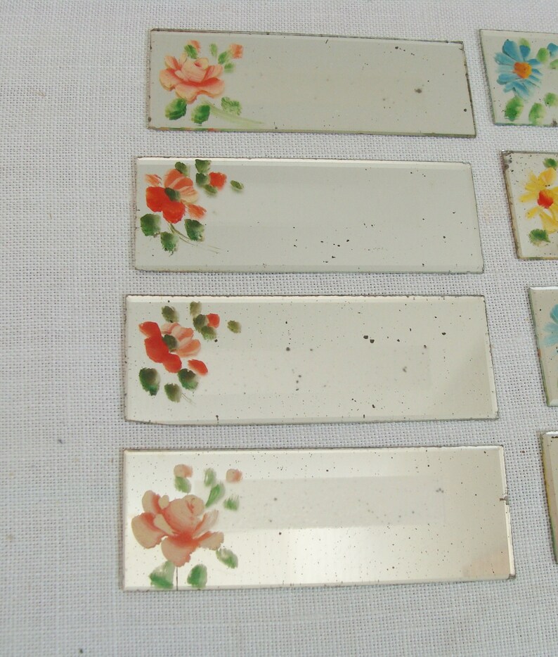 May include: Rectangular, mirrored tiles with hand-painted floral designs. The flowers are in shades of red, orange, and pink, with green leaves. The tiles are arranged on a white surface, showcasing a vintage aesthetic.