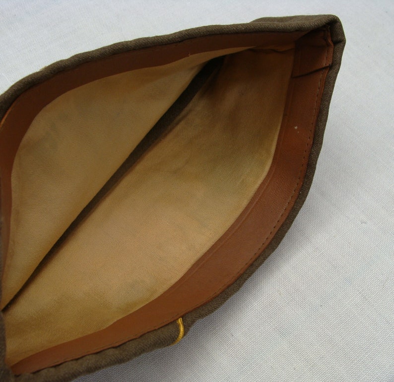 May include: An olive drab military garrison cap with a tan interior and a brown leatherette trim. The cap has a simple, folded design and a small yellow accent near the brim. The interior fabric shows signs of wear.
