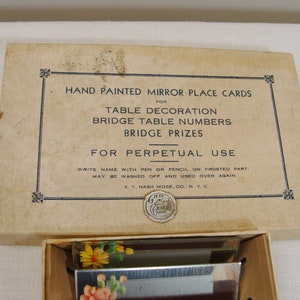 May include: A vintage box of hand-painted mirror place cards for table decoration, bridge table numbers, and bridge prizes. The box is beige with black text and a decorative border. The cards feature floral designs.
