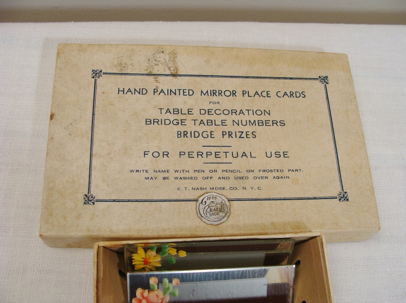 May include: A vintage box of hand-painted mirror place cards for table decoration, bridge table numbers, and bridge prizes. The box is beige with black text and a decorative border. The cards feature floral designs.