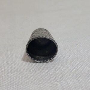 May include: A silver thimble with a detailed floral pattern around the top. The thimble has a slightly conical shape with a rounded top and a smooth, polished interior. The rim is decorated with small, evenly spaced indentations.