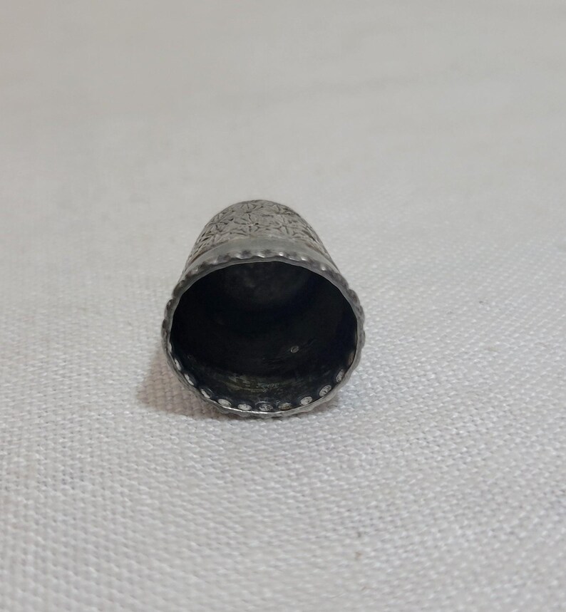 May include: A silver thimble with a detailed floral pattern around the top. The thimble has a slightly conical shape with a rounded top and a smooth, polished interior. The rim is decorated with small, evenly spaced indentations.