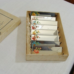 May include: A set of rectangular mirrored vanity accessories with hand-painted floral designs in a cardboard box. The flowers are in shades of red, yellow, blue, and pink. The box is a light beige color.