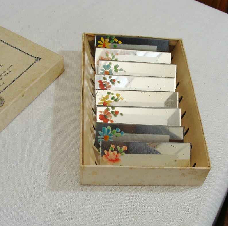 May include: A set of rectangular mirrored vanity accessories with hand-painted floral designs in a cardboard box. The flowers are in shades of red, yellow, blue, and pink. The box is a light beige color.