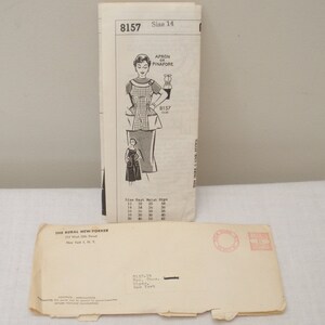 May include: Vintage sewing pattern for an apron or pinafore, size 14. The pattern features an illustration of the finished garment and includes size measurements. An envelope from The Rural New-Yorker magazine is also present.