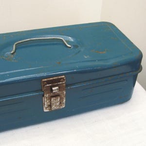 May include: A vintage teal metal toolbox with a curved handle and a rusty latch. The rectangular box shows signs of wear, with visible rust spots adding to its aged appearance. The toolbox is designed for storing tools and hardware.