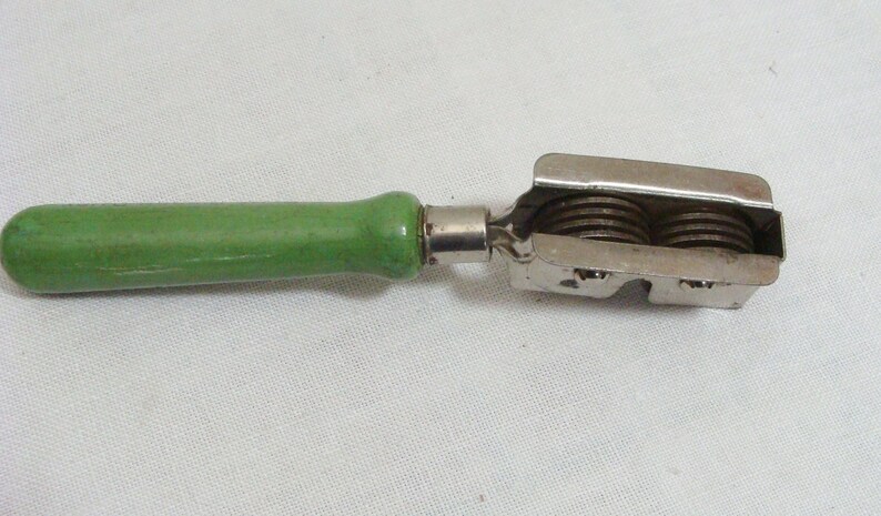May include: A vintage kitchen tool with a green wooden handle and a metal cutting head. The cutting head features two circular blades. The tool is designed for a specific food preparation task, likely slicing or cutting.