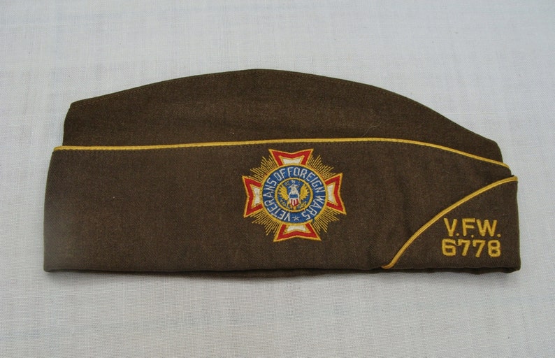 May include: A brown, triangular-shaped hat with a gold trim and embroidered emblem. The emblem reads "VETERANS OF FOREIGN WARS" and features a red, white, and blue design. The hat also has "V.F.W. 6778" embroidered in gold.