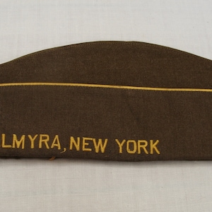 May include: A brown military-style cap with the embroidered text "PALMYRA, NEW YORK" in gold thread. The cap has a curved top and a yellow trim along the edge.