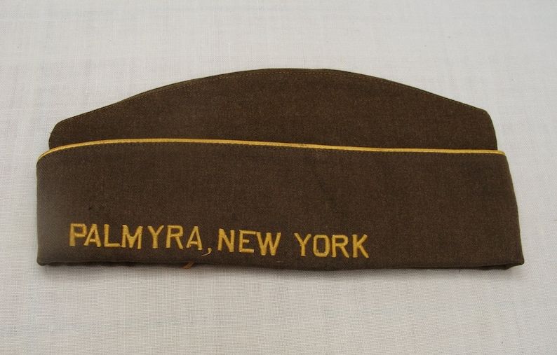 May include: A brown military-style cap with the embroidered text "PALMYRA, NEW YORK" in gold thread. The cap has a curved top and a yellow trim along the edge.