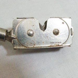May include: A vintage knife sharpener with a silver metal body and a green wooden handle. The sharpener has the text "MADE IN U.S.A." and a diamond-shaped logo with the letters "A&V".