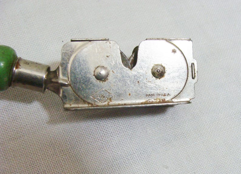 May include: A vintage knife sharpener with a silver metal body and a green wooden handle. The sharpener has the text "MADE IN U.S.A." and a diamond-shaped logo with the letters "A&V".