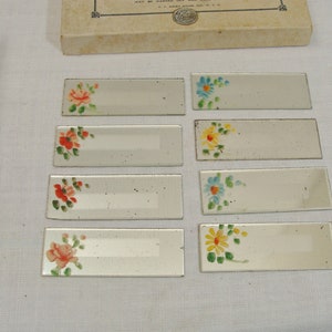 May include: Set of eight rectangular glass place card holders with hand-painted floral designs in red, orange, yellow, and blue. The place cards are arranged on a white surface, with a cardboard box in the background.