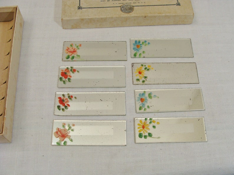 May include: Set of eight rectangular glass place card holders with hand-painted floral designs in red, orange, yellow, and blue. The place cards are arranged on a white surface, with a cardboard box in the background.