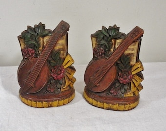 May include: Pair of decorative bookends featuring a banjo design. The bookends are brown with a yellow base and a yellow ribbon. The banjo is positioned in front of sheet music and red roses.
