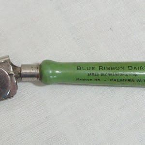 May include: Vintage can opener with a silver metal mechanism and a green handle. The handle is inscribed with "BLUE RIBBON DAIRY" and "PHONE 35 - PALMYRA, N.Y.". The can opener is designed for opening cans and is likely from the mid-20th century.