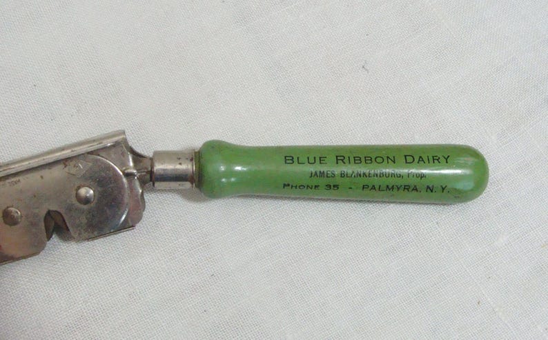 May include: Vintage can opener with a silver metal mechanism and a green handle. The handle is inscribed with "BLUE RIBBON DAIRY" and "PHONE 35 - PALMYRA, N.Y.". The can opener is designed for opening cans and is likely from the mid-20th century.