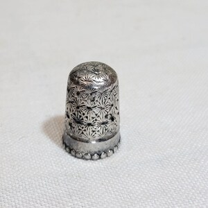 May include: A silver thimble with a detailed floral pattern. The thimble has a rounded top and a decorative beaded edge. The surface is covered in a repeating starburst design. This sewing tool is designed to protect the finger while sewing.