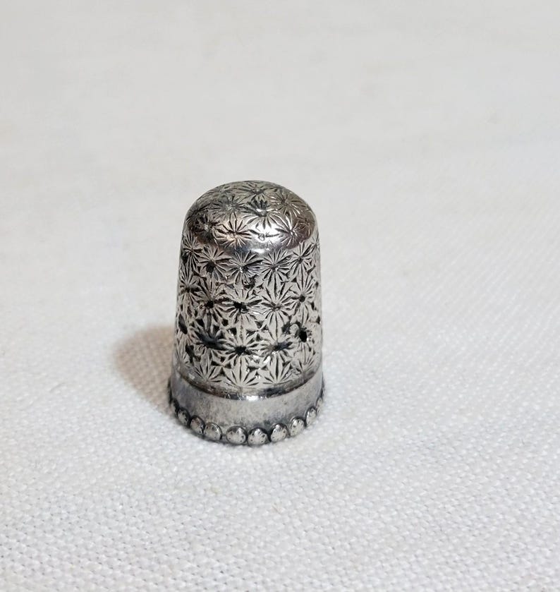 May include: A silver thimble with a detailed floral pattern. The thimble has a rounded top and a decorative beaded edge. The surface is covered in a repeating starburst design. This sewing tool is designed to protect the finger while sewing.