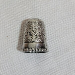 May include: A silver thimble with a detailed floral pattern. The thimble has a rounded top and a slightly flared base with a beaded edge. The surface is covered in a repeating starburst design.