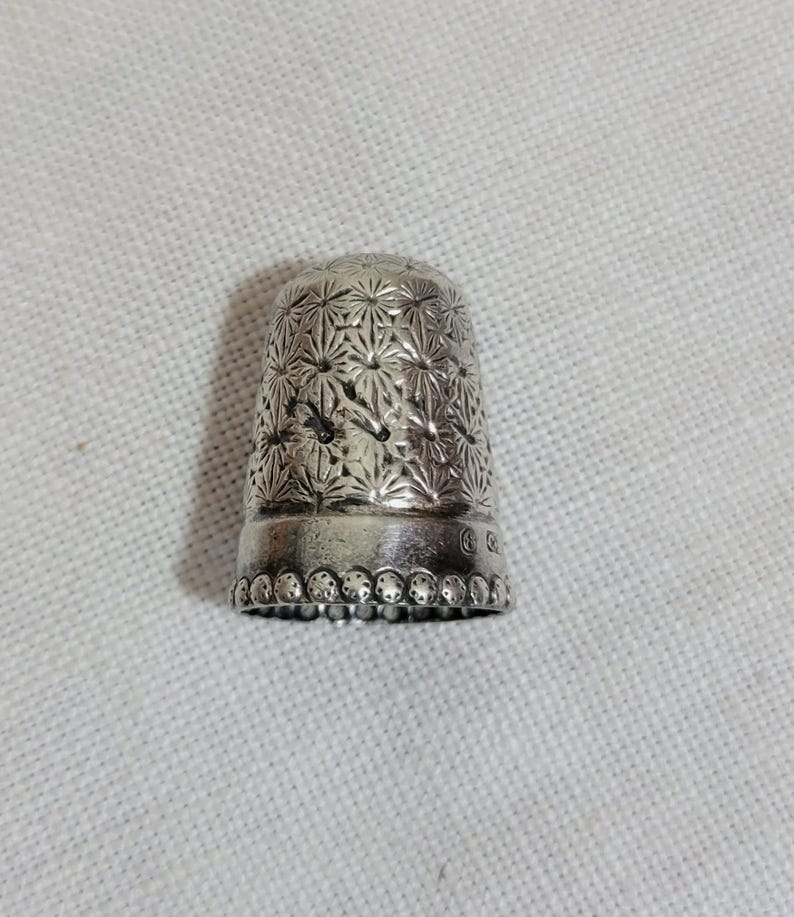 May include: A silver thimble with a detailed floral pattern. The thimble has a rounded top and a slightly flared base with a beaded edge. The surface is covered in a repeating starburst design.