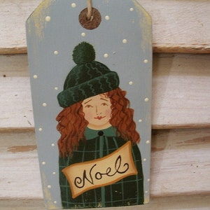 May include: A rectangular wooden gift tag painted light blue with white polka dots, featuring a hand-painted illustration of a person with curly red hair wearing a dark green knit hat and a green plaid coat, with a banner displaying the text Noel.