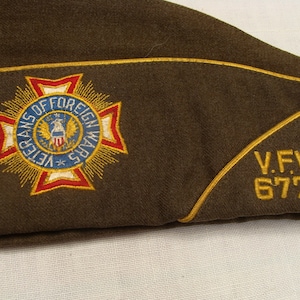 May include: A brown military garrison cap with a gold trim. The cap features an embroidered emblem of the Veterans of Foreign Wars in red, white, blue, and gold. The text "V.F.W. 6778" is embroidered in gold.