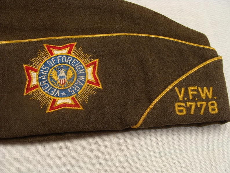 May include: A brown military garrison cap with a gold trim. The cap features an embroidered emblem of the Veterans of Foreign Wars in red, white, blue, and gold. The text "V.F.W. 6778" is embroidered in gold.