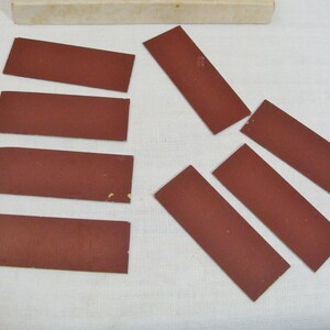 May include: A collection of rectangular, reddish-brown abrasive pads, possibly sandpaper, arranged on a white surface. A small, off-white box is visible in the background. The pads are of varying sizes and shapes.