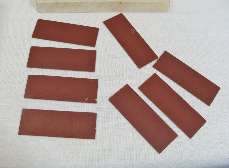 May include: A collection of rectangular, reddish-brown abrasive pads, possibly sandpaper, arranged on a white surface. A small, off-white box is visible in the background. The pads are of varying sizes and shapes.