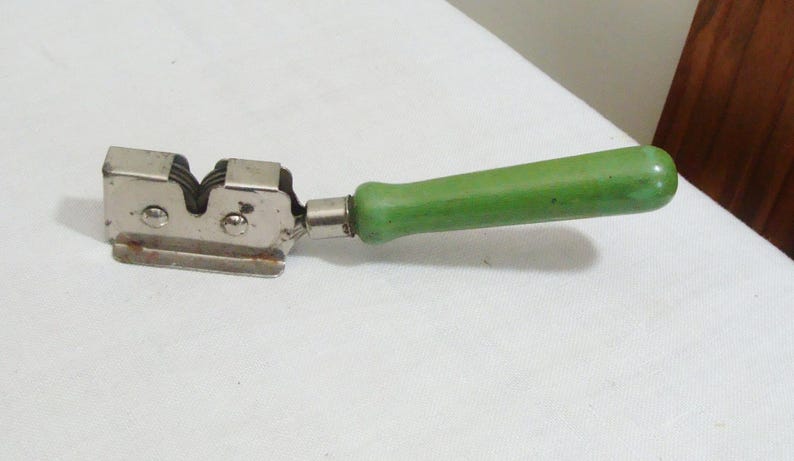 May include: A vintage knife sharpener with a silver metal body and a bright green handle. The sharpener has a double-sided design with two sharpening slots. The handle is rounded and appears to be made of wood or a similar material.