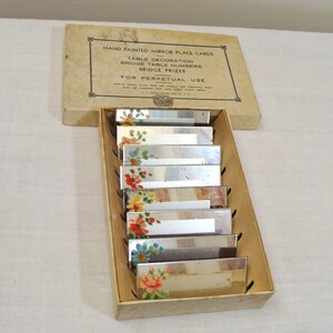 May include: A set of vintage hand-painted mirror place cards in a cardboard box. Each rectangular mirror is decorated with colorful floral designs. The box reads "HAND PAINTED MIRROR PLACE CARDS" and "FOR PERPETUAL USE".