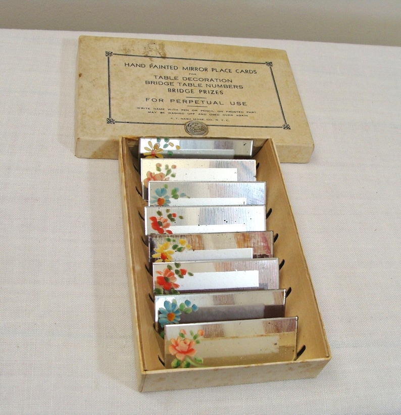 May include: A set of vintage hand-painted mirror place cards in a cardboard box. Each rectangular mirror is decorated with colorful floral designs. The box reads "HAND PAINTED MIRROR PLACE CARDS" and "FOR PERPETUAL USE".