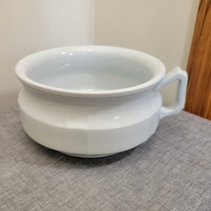 May include: A white ceramic chamber pot featuring a wide, circular opening, a smooth glazed finish, and a single angular handle on the side. The base of the vessel has a faceted, multi-sided design that tapers slightly toward the bottom rim.