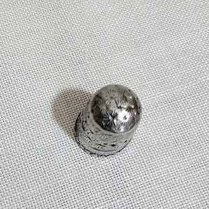 May include: A silver thimble with a detailed floral pattern. The thimble is made of metal and has a slightly rounded top. The design includes small holes around the base. This sewing tool is designed to protect the finger while sewing.