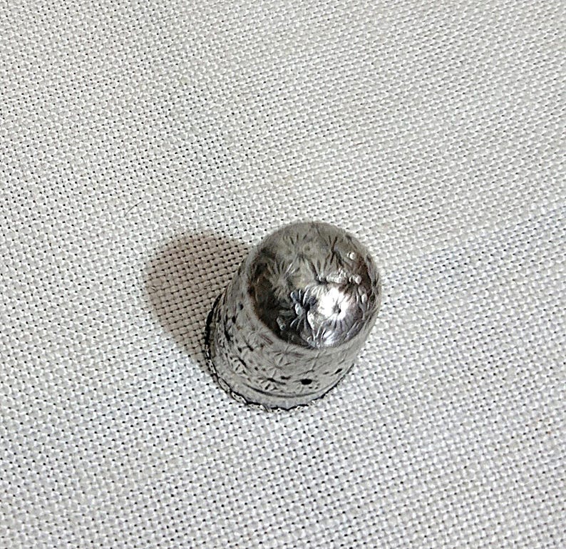 May include: A silver thimble with a detailed floral pattern. The thimble is made of metal and has a slightly rounded top. The design includes small holes around the base. This sewing tool is designed to protect the finger while sewing.