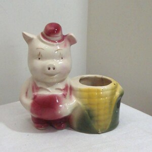 May include: A vintage ceramic pig figurine with a red hat and overalls, holding a cornucopia-shaped container. The pig is light pink with red accents, and the container is yellow and green.