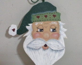 Puede incluir: Adorno navideño de madera con forma de Papá Noel. Lleva un gorro verde con corazones rojos y una barba blanca. El adorno tiene un lazo para colgar y está pintado con mejillas rosadas y una expresión sonriente. Decoración navideña.