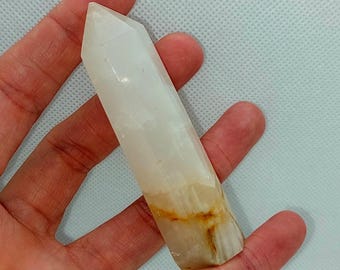 May include: A clear quartz crystal point with a polished, faceted top and a natural, rough base. The crystal has a translucent, milky white appearance with subtle amber and brown inclusions. The crystal is held in a hand.