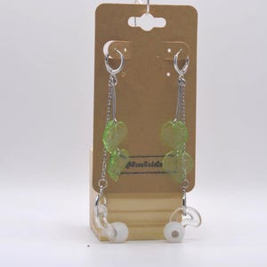 May include: A pair of silver-toned chain earrings featuring translucent green leaf-shaped charms and white earbud attachments. The earrings are displayed on a brown cardboard backing card with the text RavoBudsCo printed in black ink.