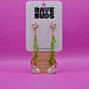 May include: A pair of gold-toned hoop earrings with dangling gold chains adorned with translucent green leaf charms and white earbud attachments. The earrings are displayed on a white card featuring the text RAVE BUDS and ravebuds.co.