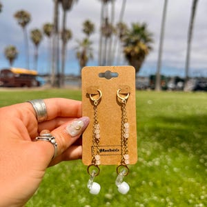 May include: A pair of gold-toned dangle earrings featuring thin chains, small pale pink beads, and white earbud-style attachments. The earrings are displayed on a rectangular brown cardboard card with the text @RaveBudsCo printed in black ink.
