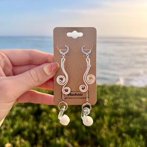 May include: A pair of silver-toned wire earrings featuring a double-spiral design attached to lever-back hooks. Each earring includes a small silver chain connecting to a clear, circular plastic link and a white, rounded earplug-style charm.