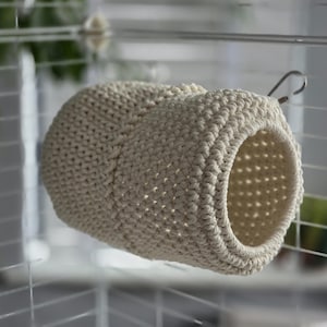 May include: A beige, crocheted pet hideaway, shaped like a small tunnel, hangs inside a wire cage. The handmade item features a circular opening and a sturdy, textured design. The neutral colour and open weave provide a cosy space for small animals.