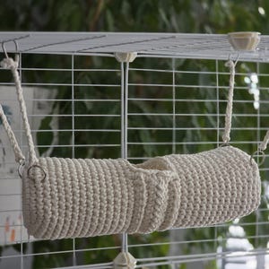 May include: A cream-coloured, crocheted pet hammock suspended from a wire cage. The hammock is cylindrical with a central opening and is attached by braided rope and metal rings. The background is a blurred view of greenery.