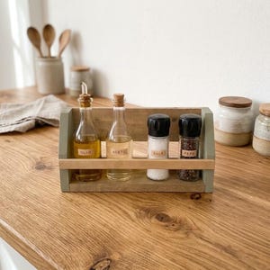 Pode incluir: Um suporte de madeira para condimentos com quatro recipientes de vidro: duas garrafas transparentes com rolhas de cortiça para azeite e vinagre, e dois saleiros com tampas pretas para sal e pimenta. Cada recipiente tem um rótulo: Olio, Aceto, Sale, Pepe.